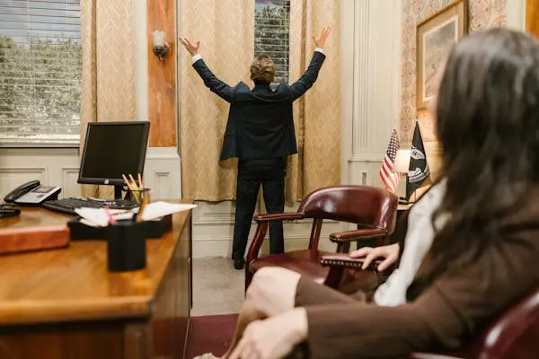 A lawyer stands facing the window with hands raised, while a colleague sits nearby in a law office, reflecting a moment of legal strategy and contemplation.