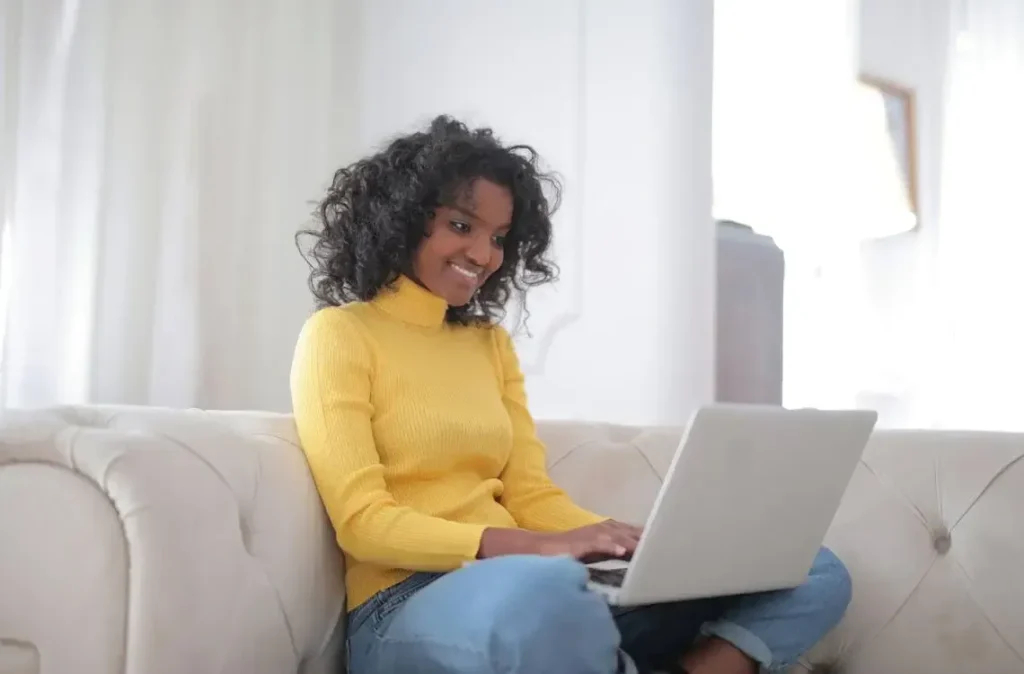 Smiling young ethnic lady working on her laptop while sitting at couch at home symbolizing confidence.